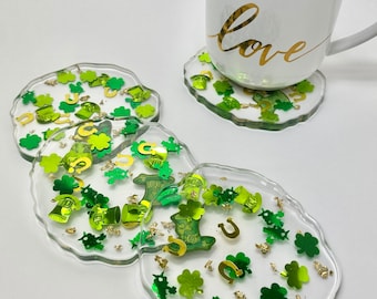 May include: Four clear resin coasters with a green and gold shamrock, horseshoe, and leprechaun hat design. The coasters are on a white surface with a white mug that says "love" on it.