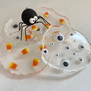 May include: Clear resin coasters with candy corn and googly eyes. A black glitter spider sits atop the coasters. Halloween-themed home decor.