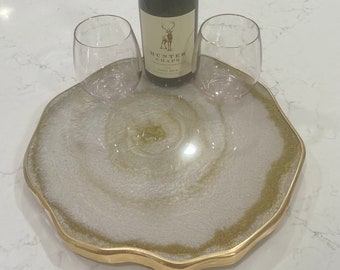 May include: A white and gold resin serving tray with a wavy edge. The tray has a swirling pattern in the center. Two wine glasses and a bottle of Hunter Chaps Pinot Noir wine are on the tray.