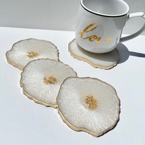 May include: Four white coasters with gold edges and gold flecks. The coasters are arranged in a stack with the top coaster slightly offset. A white mug with gold lettering that says "Love" is sitting on top of the coasters.