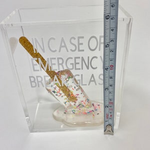 May include: A clear acrylic box with a white background. Inside the box is a white popsicle with gold glitter and colourful sprinkles. The popsicle is melting and dripping down the side of the box. The box has the text "IN CASE OF EMERGENCY BREAK GLASS" printed on the side.