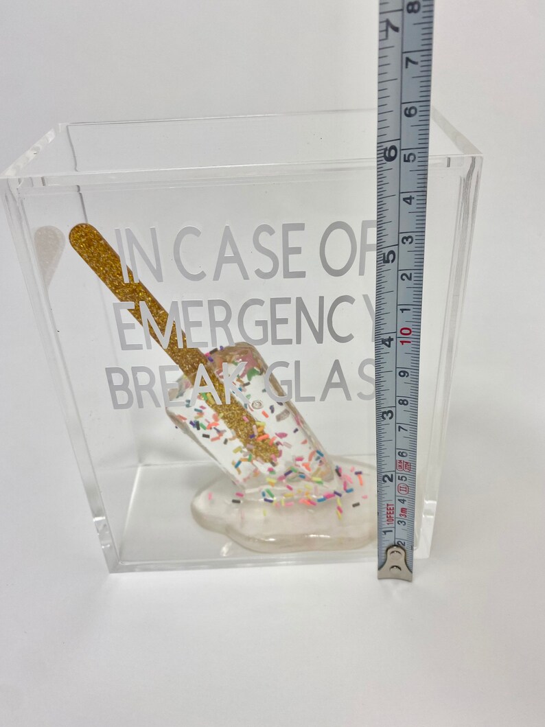 May include: A clear acrylic box with a white background. Inside the box is a white popsicle with gold glitter and colourful sprinkles. The popsicle is melting and dripping down the side of the box. The box has the text "IN CASE OF EMERGENCY BREAK GLASS" printed on the side.