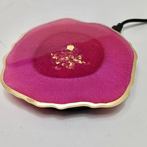 Peut inclure: Un chargeur sans fil rose et or avec un bord ondulé. Le chargeur est en résine et présente un motif en feuille d'or.