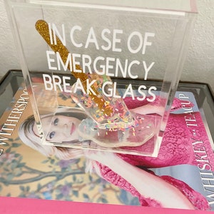 May include: A clear acrylic box with the text "In case of emergency break glass" printed on the front. Inside the box is a gold glitter-covered glass rod with colourful sprinkles.