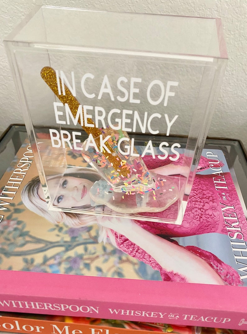 May include: A clear acrylic box with the text "In case of emergency break glass" printed on the front. Inside the box is a gold glitter-covered glass rod with colourful sprinkles.