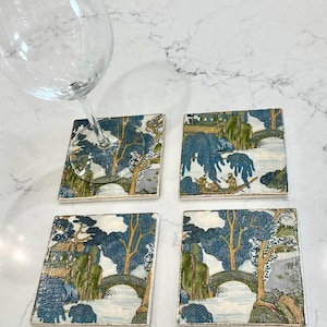 Op de afbeelding: Set van vier vierkante keramische onderzetters met een blauw en groen landschapsontwerp. Elke onderzetter heeft een brug, bomen en een rivierlandschap. Een wijnglas rust op een van de onderzetters.