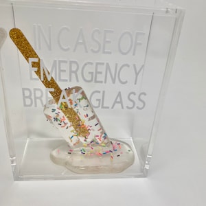 May include: A clear acrylic box containing a white popsicle. The popsicle is covered in colourful sprinkles and has a gold glitter stick. The text "IN CASE OF EMERGENCY BREAK GLASS" is printed on the box.