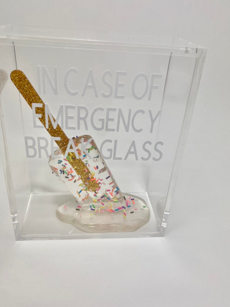 May include: A clear acrylic box containing a white popsicle. The popsicle is covered in colourful sprinkles and has a gold glitter stick. The text "IN CASE OF EMERGENCY BREAK GLASS" is printed on the box.