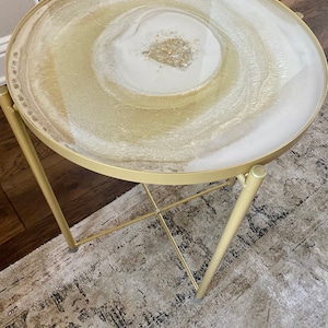 Puede incluir: Una mesa auxiliar de metal dorado con una tapa redonda de resina blanca y dorada. La tapa tiene un patrón de remolino y detalles de purpurina dorada.