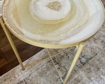 May include: A gold metal side table with a round, white and gold resin top. The top has a swirling pattern and gold glitter accents.