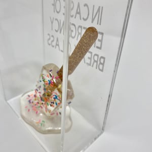 May include: A clear acrylic box containing a white, melted, and glittery object. The object is covered in colourful sprinkles. The box has the text "BREAK GLASS IN CASE OF EMERGENCY" printed on the side.