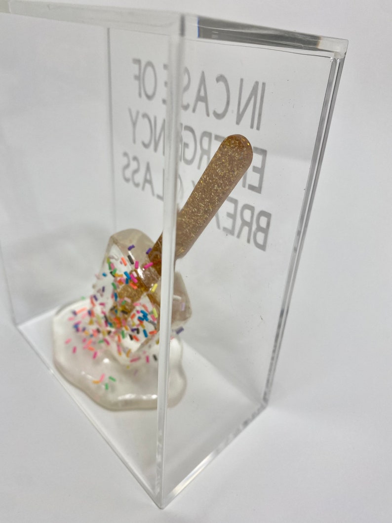 May include: A clear acrylic box containing a white, melted, and glittery object. The object is covered in colourful sprinkles. The box has the text "BREAK GLASS IN CASE OF EMERGENCY" printed on the side.