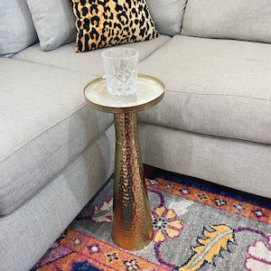May include: A gold metal side table with a hammered finish and a round top. The table has a glass on top and is sitting on a colourful rug.
