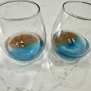 May include: Two clear glass wine glasses containing a miniature beach scene. Each glass features brown sand, blue water, and tiny figures of people.