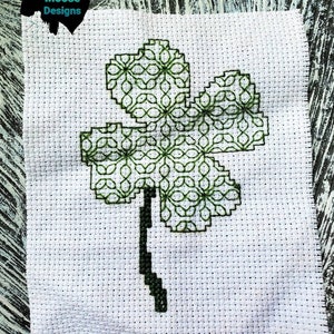 May include: A green four-leaf clover cross-stitch pattern on white Aida cloth. The clover is outlined in black and filled with a green geometric pattern.