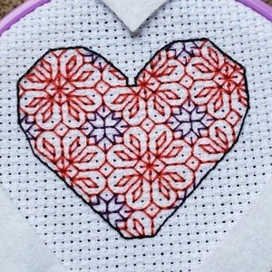 May include: A white embroidery hoop with a purple outer ring, featuring a red and purple heart-shaped cross-stitch design.