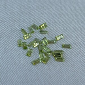 Può includere: Una collezione di pietre preziose rettangolari di colore verde chiaro, note anche come peridoto, sparse su uno sfondo di tessuto bianco.