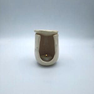 Puede incluir: Un portavelas de cerámica color crema con una pequeña vela encendida en su interior. El soporte tiene una abertura en forma de U y un diseño orgánico texturizado. La parte superior tiene una pequeña tapa plana.