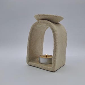 May include: An arched beige ceramic oil burner with a textured, matte finish. The structure features a top basin for oil and a hollow centre space containing a small, round metal tea light candle holder with a lit candle inside the burner.