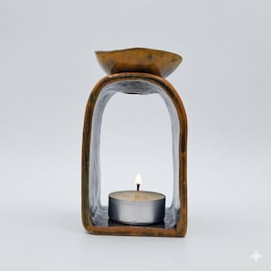 May include: A brown ceramic oil burner with a tall, arched frame and a shallow bowl at the top. A small, round tea light candle with a lit flame sits centred on the base within the arch, casting a warm glow against the smooth, glazed surface.