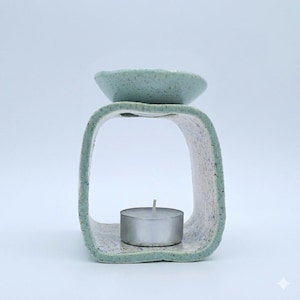 May include: A ceramic oil burner with a speckled light blue and white finish. The structure features a square-shaped frame with rounded corners and a small bowl at the top. A single silver tea light candle sits inside the base of the frame.