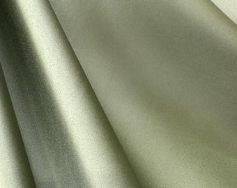 May include: Close-up of a folded fabric in a soft, muted green color. The fabric has a subtle sheen and a smooth texture, with visible folds and shadows creating depth. The overall appearance suggests a luxurious textile.