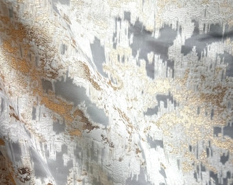 May include: White fabric with a gold metallic abstract pattern. The fabric has a subtle sheen and a textured surface.