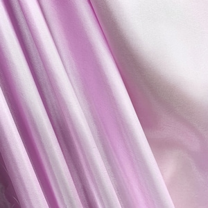 May include: A close-up of a soft, silky fabric in a light purple color. The fabric is draped and folded, creating a smooth, flowing texture.