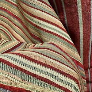 May include: Close-up of a textured fabric with a striped pattern. The fabric features alternating stripes of red, green, tan, and gray. The fabric is folded, creating a sense of depth and texture. The overall theme is home decor.