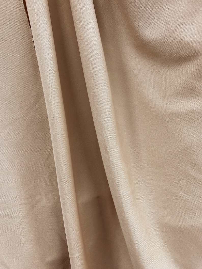 May include: A close-up of a smooth, beige fabric with a subtle sheen. The fabric is folded in a way that creates soft, flowing lines.