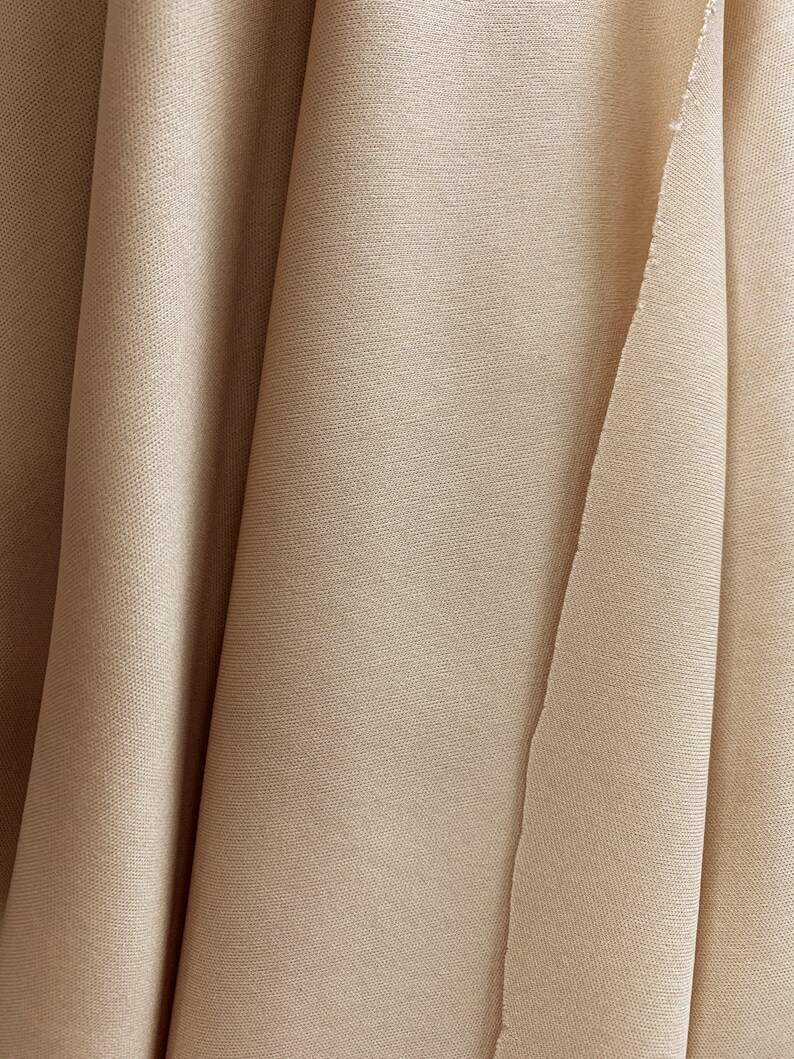 May include: A close-up of a beige fabric with a smooth texture. The fabric is folded in a way that creates a series of soft curves.
