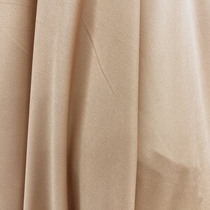 May include: Close-up of a smooth, light brown fabric with subtle folds and wrinkles. The fabric has a soft, silky texture.
