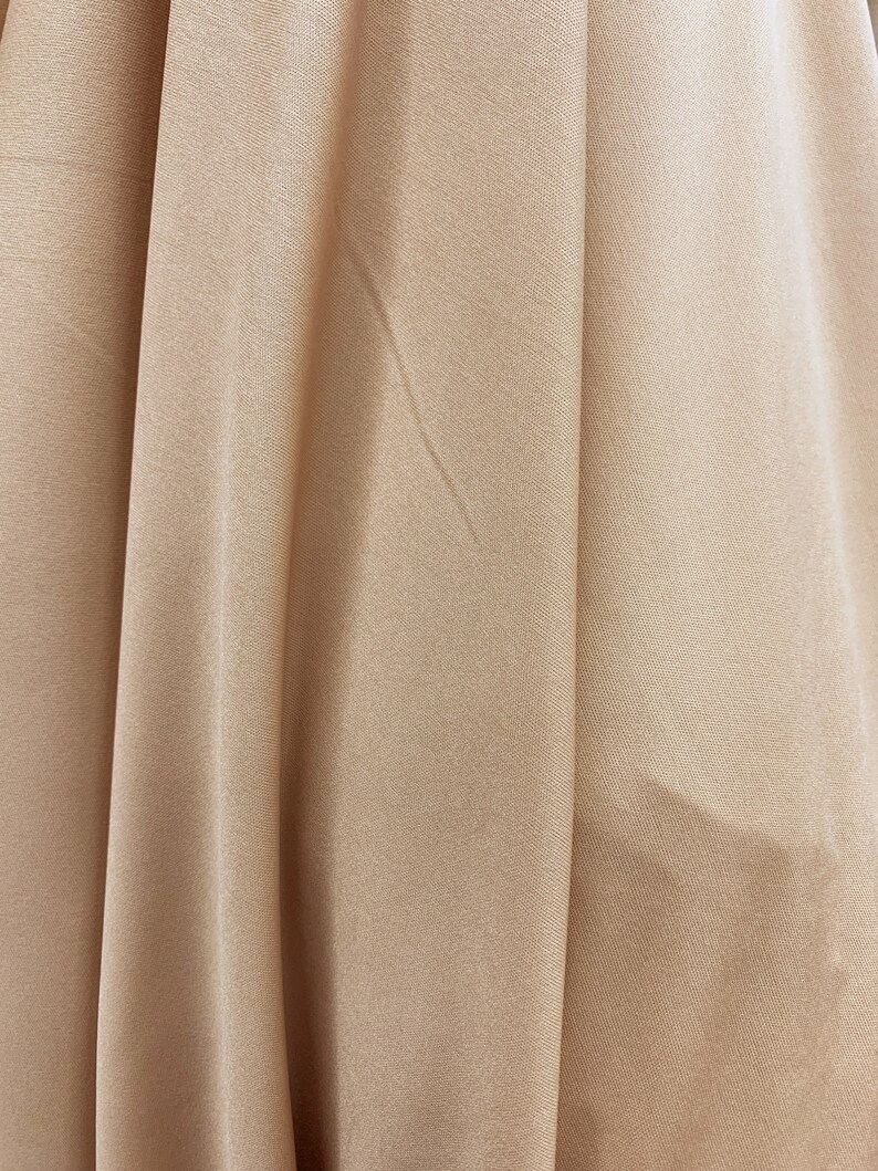 May include: Close-up of a smooth, light brown fabric with subtle folds and wrinkles. The fabric has a soft, silky texture.