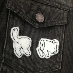 Peut inclure: Gros plan sur une veste en jean gris foncé avec deux écussons en feutre blanc. L'un représente l'arrière d'un animal de dessin animé, l'autre une tête d'animal de dessin animé. La veste a une poche et un bouton argenté.