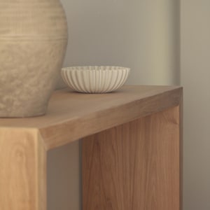 Op de afbeelding: Een lichtbeige keramische kom met een gegroefd, geribbeld exterieurontwerp staat op een houten consoletafel naast een grote, getextureerde, lichtbruine keramische vaas. De kom heeft een ronde vorm en een consistent, verticaal patroon.