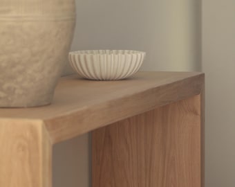 Könnte beinhalten: Eine hellbeige Keramikschale mit geriffeltem Außendesign steht auf einem Holzkonsolentisch neben einer großen, strukturierten, hellbraunen Keramikvase. Die Schale hat eine abgerundete Form und ein gleichmäßiges, vertikales Muster.