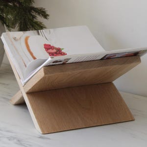 Può includere: Un leggio in legno per libri di cucina con base a forma di X, che tiene un libro di cucina aperto. Il leggio è in legno chiaro e ha una finitura naturale. Il libro di cucina mostra una foto di un dolce con frutti rossi.