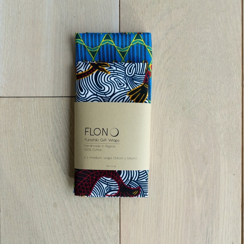 May include: Two blue and white fabric gift wraps with geometric and abstract patterns. The wraps are made in Nigeria from 100% cotton and measure 22 inches by 22 inches. The packaging includes the brand name "FLONO" and the text "Furoshiki Gift Wraps".