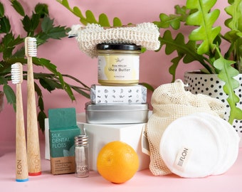 Puede incluir: Una colección de productos de baño de cero residuos, que incluye un tarro de manteca de karité africana cruda, una pastilla de jabón, una lata de metal, dos cepillos de dientes de bambú, un pequeño frasco de vidrio de hilo dental de seda, un disco de algodón reutilizable, una bolsa de malla para productos y una naranja.