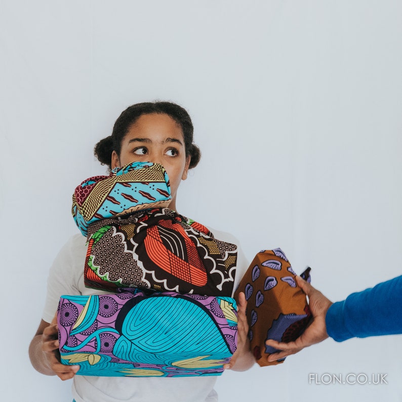 May include: A person holding a stack of colorful fabric-wrapped gifts. The gifts are decorated with geometric patterns and bright colors. The person is wearing a white shirt and is looking to the right.