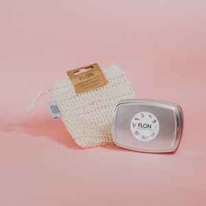 Pode incluir: Uma bolsa de malha branca com uma etiqueta marrom que diz "FLON" e um recipiente de metal prateado com uma etiqueta branca que diz "FLON" sobre um fundo rosa.