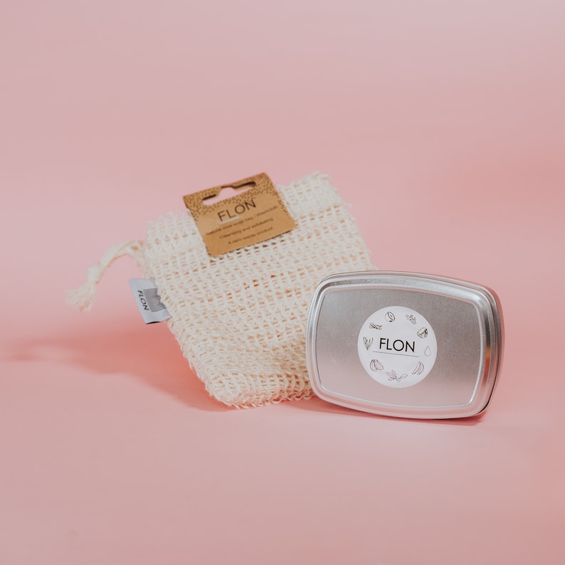 Pode incluir: Uma bolsa de malha branca com uma etiqueta marrom que diz "FLON" e um recipiente de metal prateado com uma etiqueta branca que diz "FLON" sobre um fundo rosa.