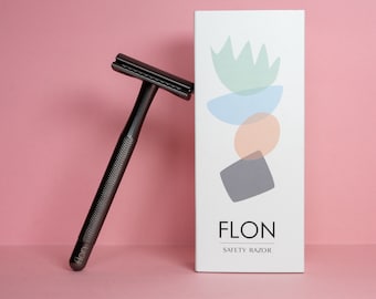 以下が含まれることがあります： ハンドルに「FLON」と印刷された黒い安全かみそり。かみそりは、「FLON SAFETY RAZOR」と印刷された白い箱の隣にあります。箱にはカラフルな抽象的なデザインが施されています。
