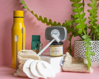 Puede incluir: Una colección de productos de baño de cero residuos sobre una superficie rosa. Los productos incluyen una botella de agua de metal amarilla, una pequeña caja verde etiquetada como "Silk Dental Floss", un pequeño contenedor de metal, una barra de manteca de karité blanca etiquetada como "Raw African Shea Butter", una lata de metal blanca con la palabra "FLON" en ella, una cuchilla de afeitar blanca, dos cepillos de dientes de madera y una bolsa de malla blanca con discos de algodón blancos.