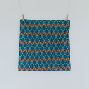 May include: A blue fabric with a repeating pattern of red, yellow, and green geometric shapes. The fabric is hanging from two clothespins.
