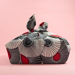 May include: A black and white fabric with red accents, folded into a square shape with a knot at the top. The fabric has a geometric pattern of overlapping circles and lines.