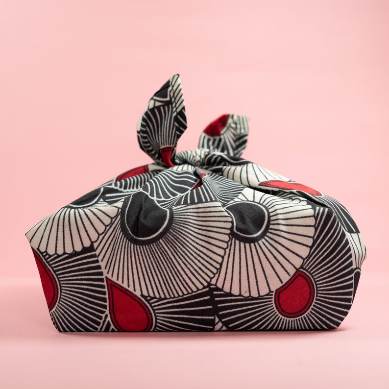 May include: A black and white fabric with red accents, folded into a square shape with a knot at the top. The fabric has a geometric pattern of overlapping circles and lines.