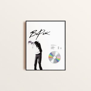 May include: A framed poster with a black and white photo of a person in a white shirt and black pants. The poster has the text "BLACKPINK" in black cursive writing at the top. A silver CD with a rainbow and pink design is in the bottom right corner of the poster.