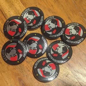 May include: Seven black and white round buttons with the words "ACCIÓN ANTIFASCISTA" in white lettering. The buttons have a red and white graphic design.