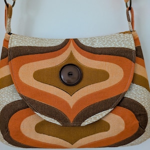 May include: A shoulder bag featuring a repeating geometric pattern in shades of orange, brown, and cream. The bag has a flap closure secured by a single large, dark brown circular button. It includes a fabric strap attached with metal rings.
